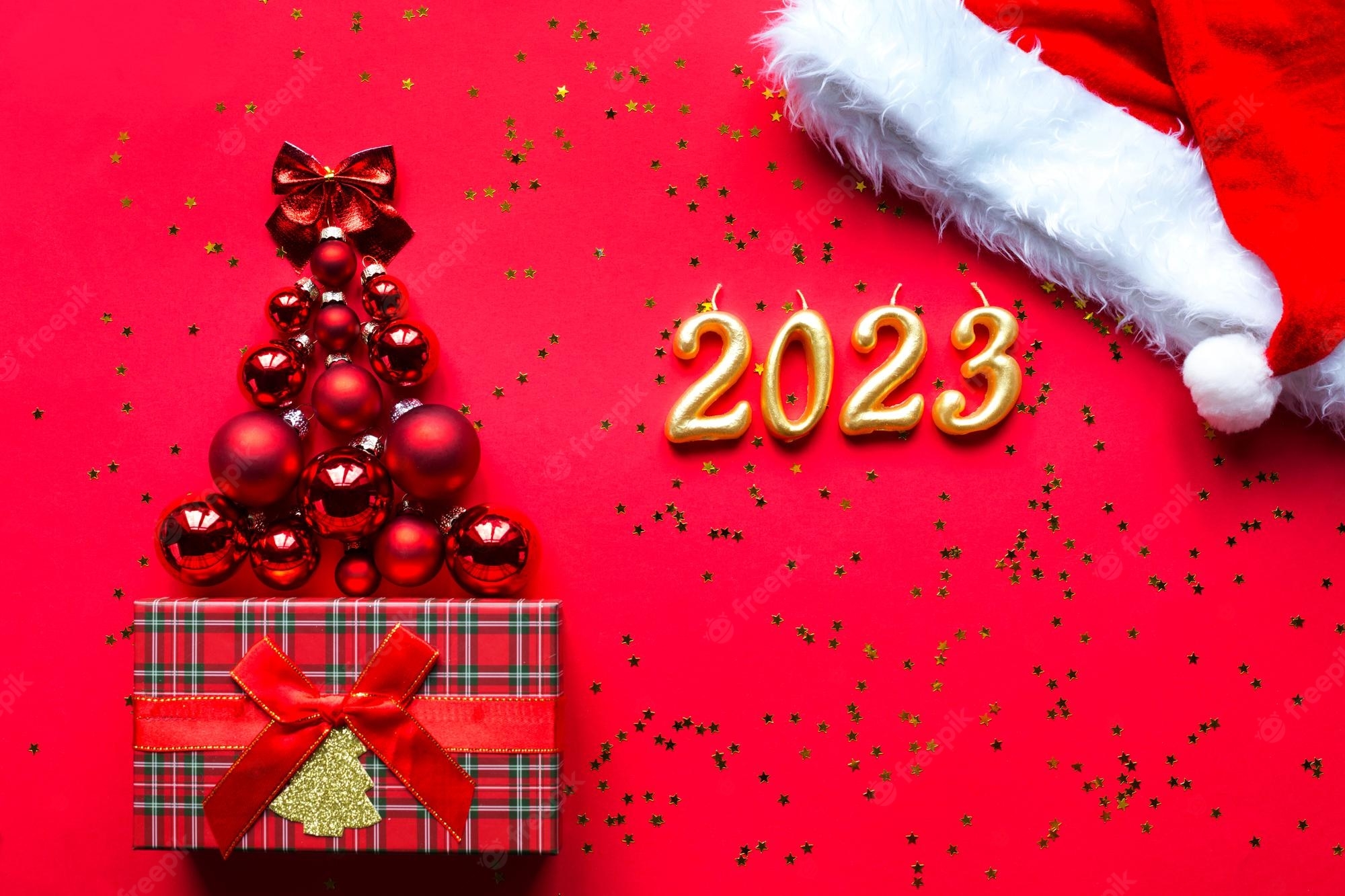 2000x1340 Premium Photo. Christmas tree made of glass balls and the numbers 2023 from candles on a red background with gold stars sequins. christmas background, new year, santa hat and gift box, Desktop